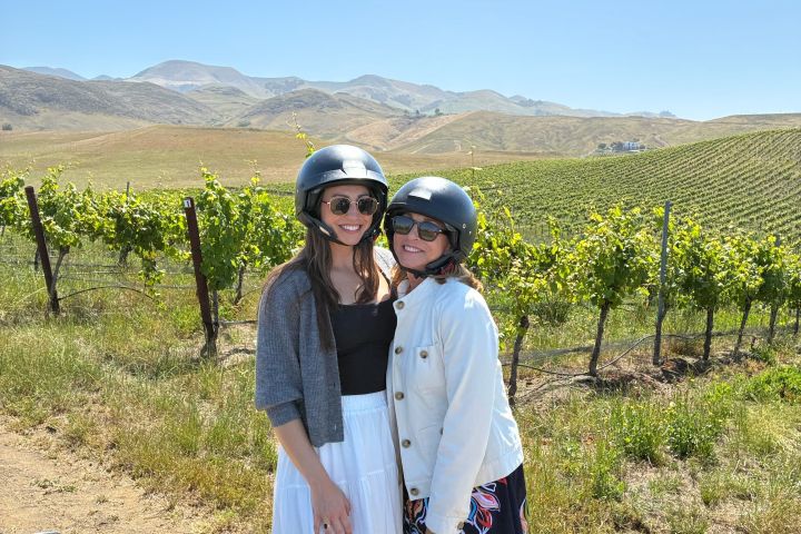Two people in helmets stand smiling in a vineyard with hills in the background.