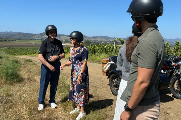 Group of people wearing helmets standing by motorcycles in a grassy field.